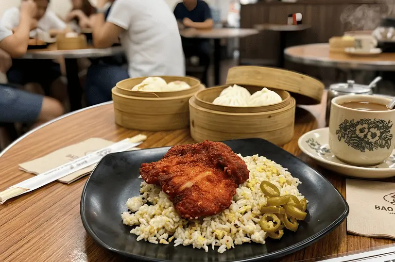 A plate of fried rice topped with a large piece of crispy fried chicken and pickled green chilies, accompanied by bamboo steamer baskets of dim sum and a traditional floral coffee cup.