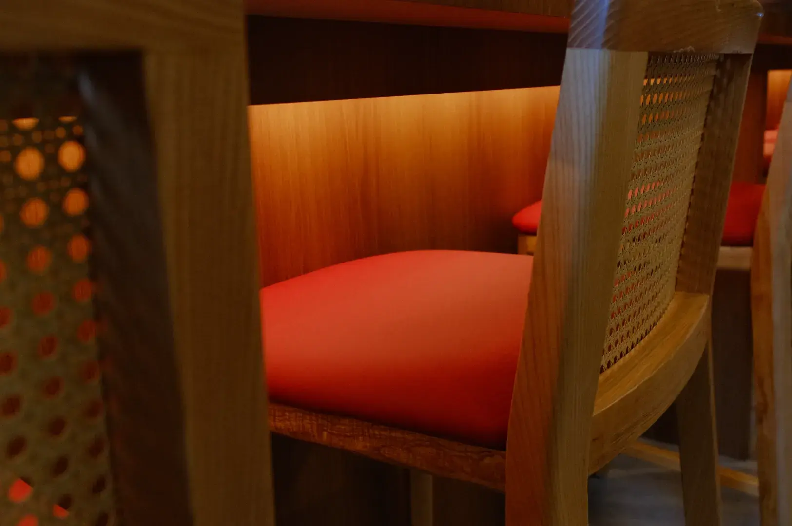 A close-up view of wooden chairs with red cushions, situated around a bar or counter with soft warm lighting.