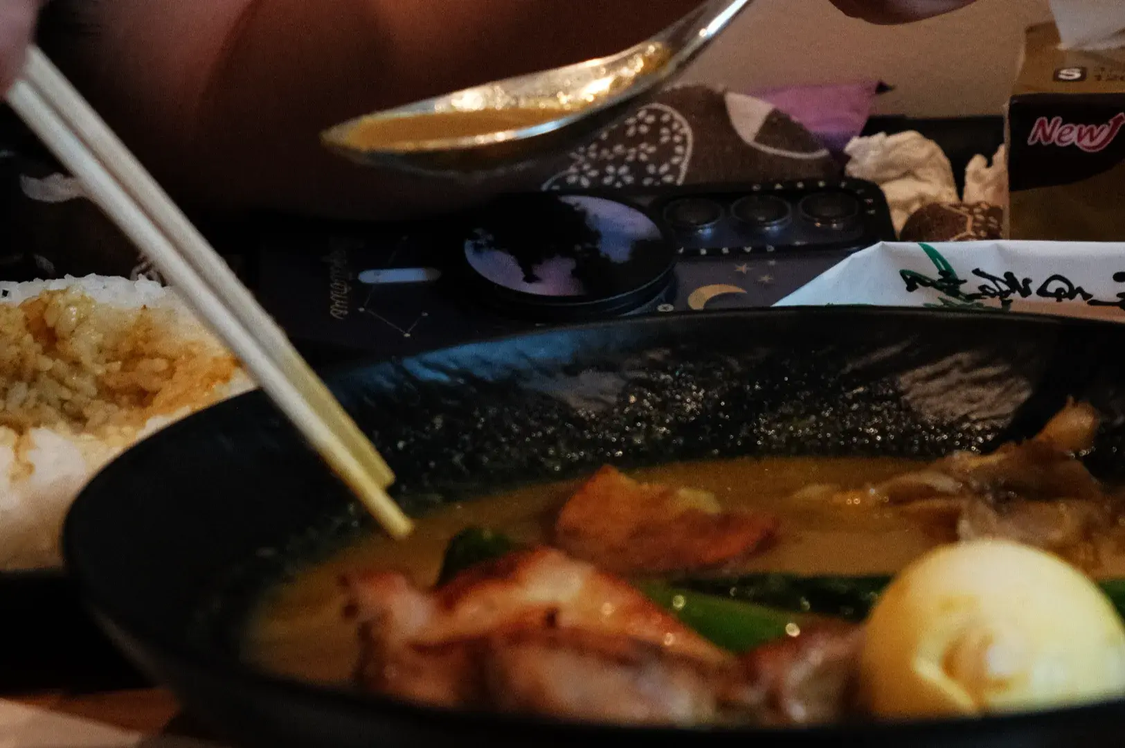 A bowl of soup curry with chicken and vegetables, with chopsticks and a spoon nearby, and a plate of rice in the background.