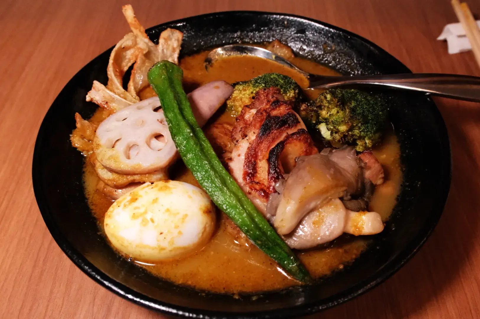 A bowl of ramen with various toppings including sliced meats, a soft-boiled egg, broccoli, lotus root, and a long green vegetable, served in a rich broth.
