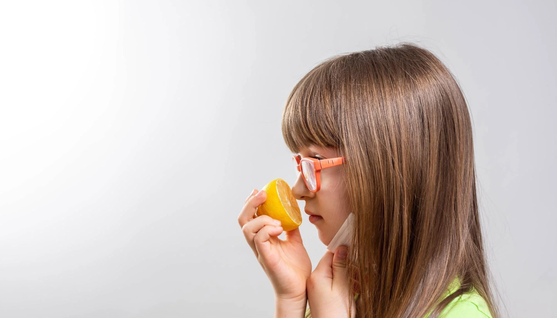 «Jente med langt brunt hår og oransje briller holder en halv sitron nær nesen mens hun tar av seg munnbindet.»
