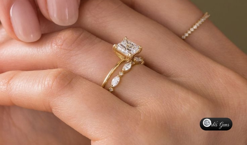 A delicate hand displays a stunning gold ring featuring a Princess Cut Diamond, accentuated by smaller marquise and round diamonds along the band, showcasing the timeless beauty of Princess Cut Diamonds.