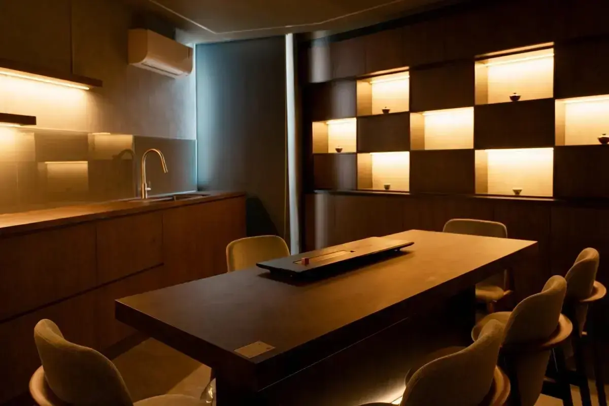 This interior features a minimalist dining or tea room characterized by dark wood cabinetry, warm recessed lighting, and a checkered display wall showcasing small vessels. A long, solid table anchors the space, surrounded by curved chairs and set against a sleek kitchenette with a gold-toned faucet.