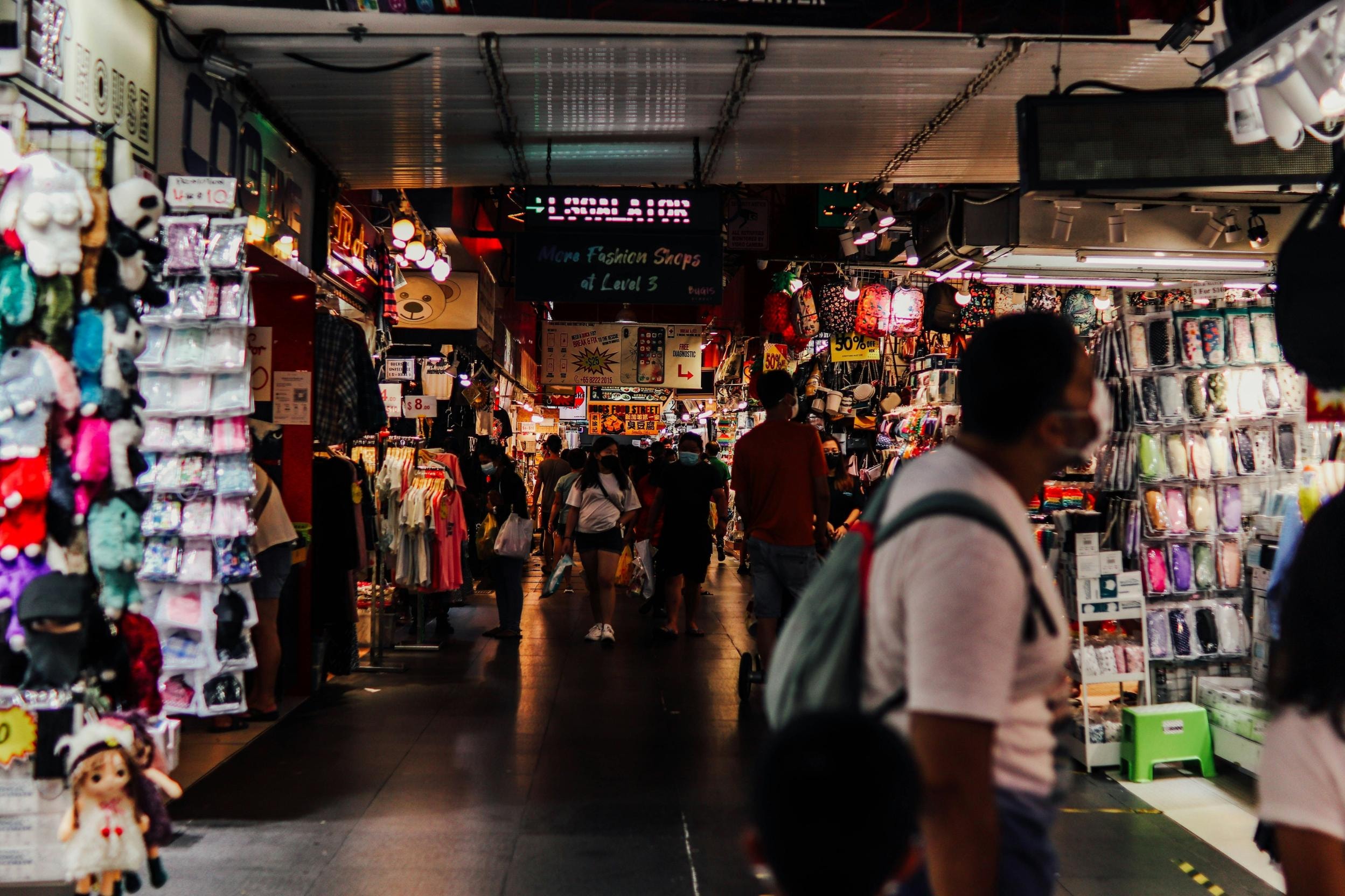 This image depicts a crowded and bustling indoor market corridor, where narrow aisles are tightly packed with stalls displaying a diverse array of merchandise like plush toys, clothing, and accessories. Shoppers navigate the dimly lit walkway under the glow of neon signage, creating a vibrant and chaotic retail atmosphere typical of busy street markets.