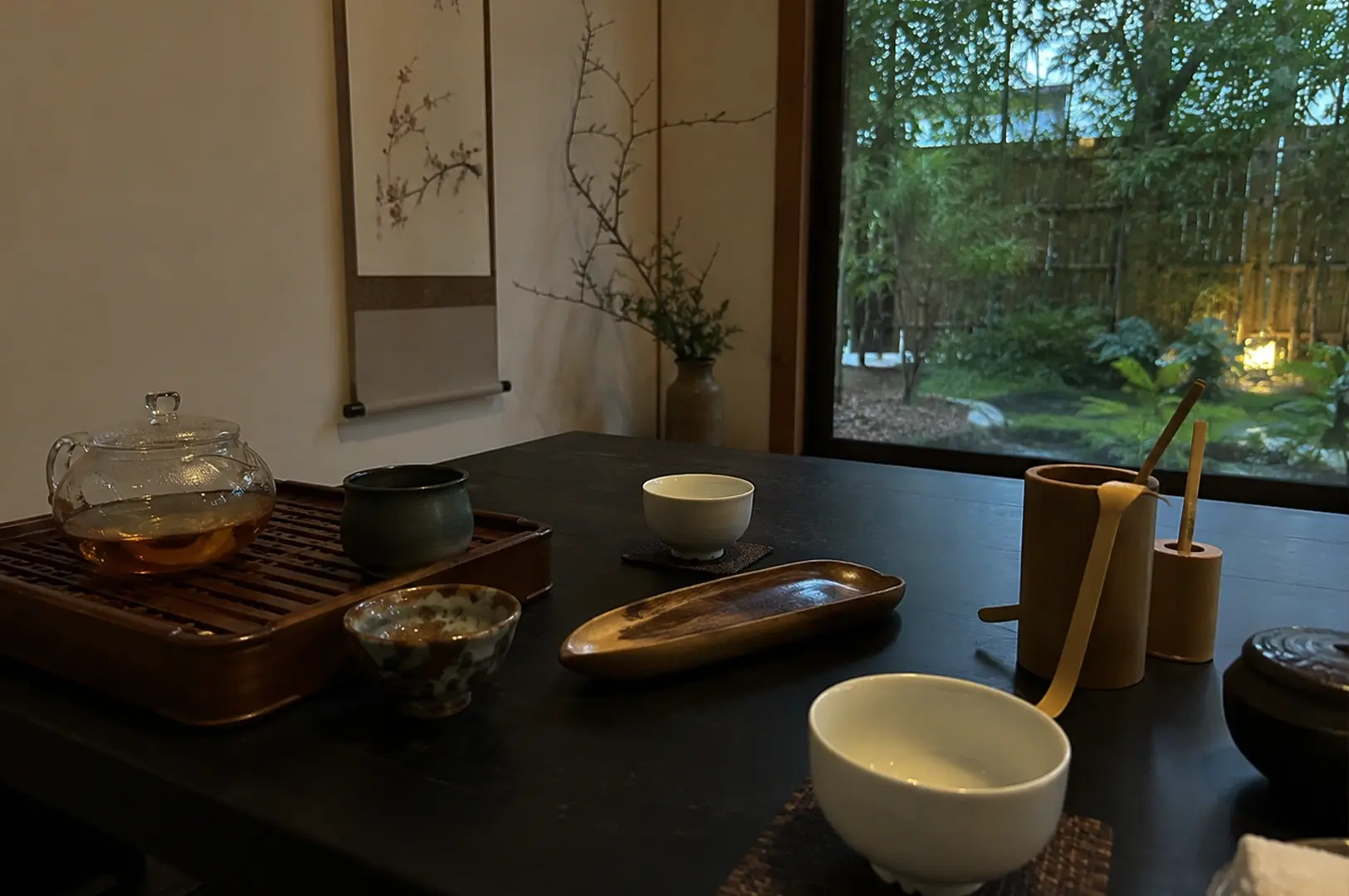 A serene tea setting is arranged on a dark, rustic wooden table next to a large window looking out onto a lush, illuminated bamboo garden at dusk. The arrangement features an assortment of tea-making utensils, including a clear glass teapot filled with amber liquid on a wooden slatted tray, a long carved wooden leaf-shaped tea scoop container, bamboo tea scoops resting upright in a holder, and small ceramic teacups placed on textured fabric coasters. The peaceful atmosphere is enhanced by a minimalist hanging scroll featuring delicate floral calligraphy and a simple branch arrangement in a tall ceramic vase against the muted wall.