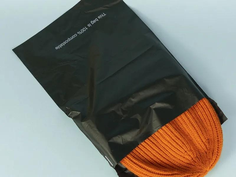 A black mailer partially opened, with a beanie hat peeking out. The beanie appears soft and cozy, suggesting it is made of knitted or wool material. The black mailer has a sleek and durable design, commonly used for shipping clothing and accessories. 