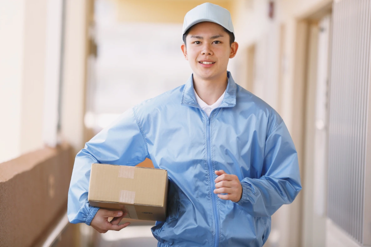 A person in a light blue jacket and cap holds a cardboard box while walking in a hallway.