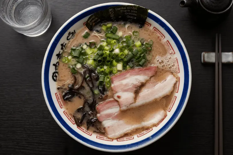 A bowl of ramen with slices of pork, green onions, and seaweed on a wooden table. The broth appears rich and creamy, creating a cozy atmosphere.