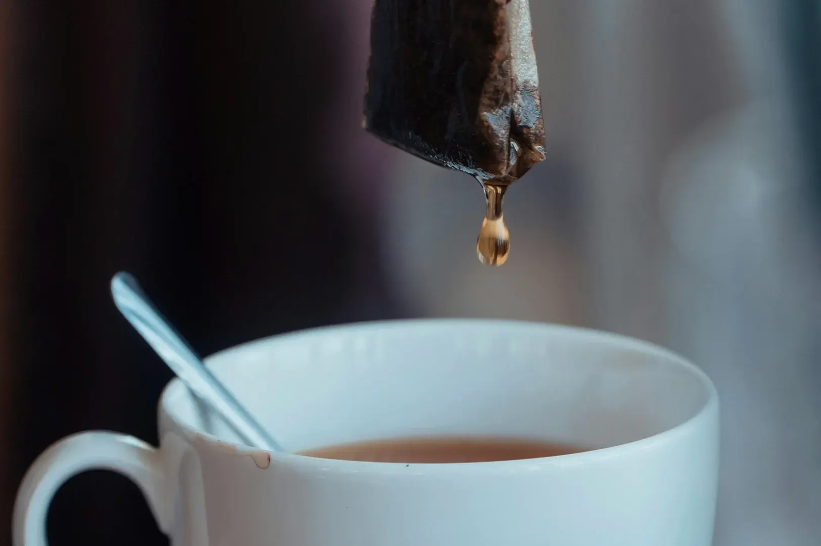 A tight macro shot captures a wet, brown tea bag suspended in mid-air, frozen just as a single, translucent amber droplet of tea hangs from its bottom corner. Below, a portion of a clean white ceramic mug is visible, showing a silver metal spoon resting inside against the rim and a hint of the steeped tea liquid surface, all set against a soft, heavily blurred background of dark and muted grey tones.