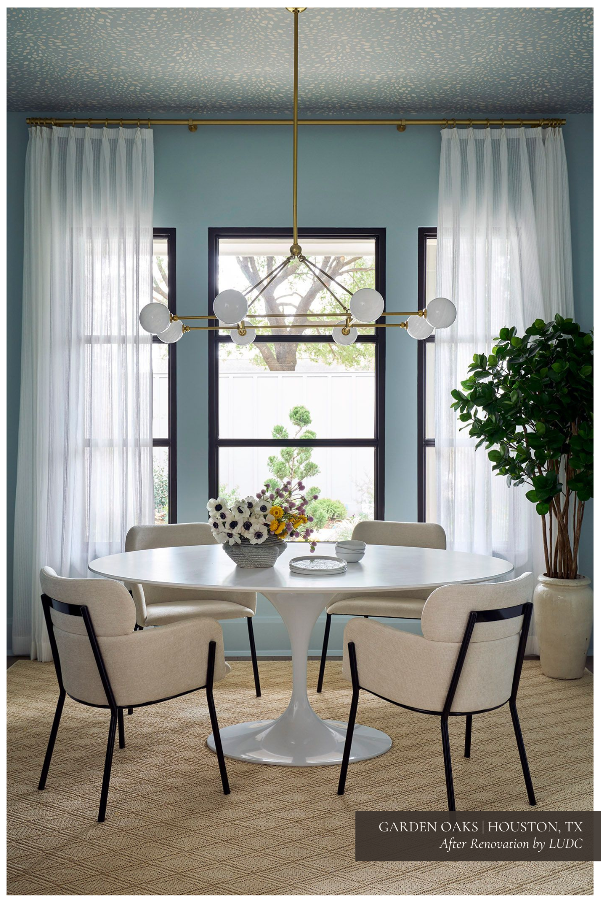 A playful breakfast nook featuring a Rove Concepts Tulip table, modern chairs, a textured sisal rug, and a wallpapered ceiling, with a brass chandelier adding a touch of glamour.