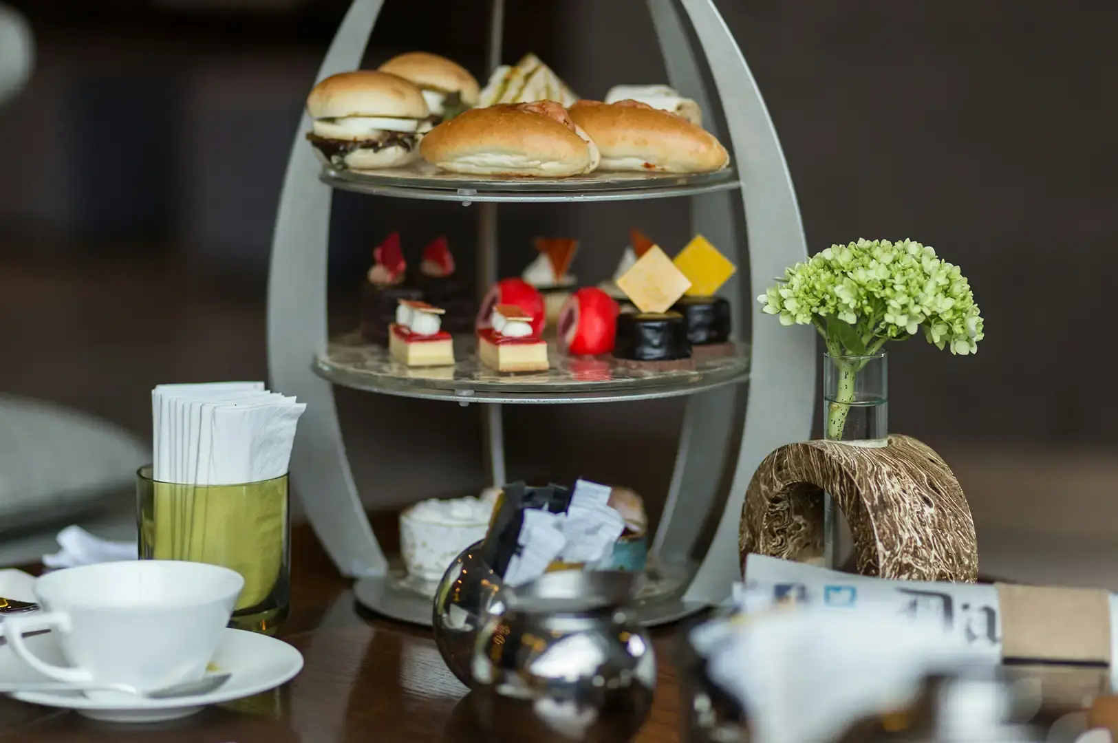 A sleek, oval-framed three-tiered afternoon tea stand sits on a dark wooden table, holding mini slider sandwiches and stuffed buns on the top tier, colorful layered mousse pastries and chocolate bites on the middle tier, and cream desserts on the bottom tier, set alongside a white teacup, a glass utensil holder, and a small wooden vase holding green hydrangeas.