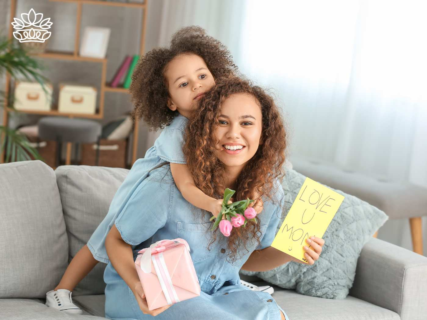 A mother joyfully holding a "Love U Mom" card, pink gift box, and tulips while embracing her daughter, representing the Gift Boxes for Her Collection - Fabulous Flowers and Gifts