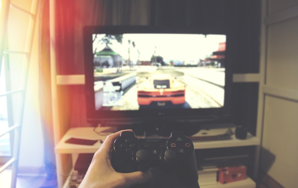 A hand holding a video game controller with a blurred out TV in the background