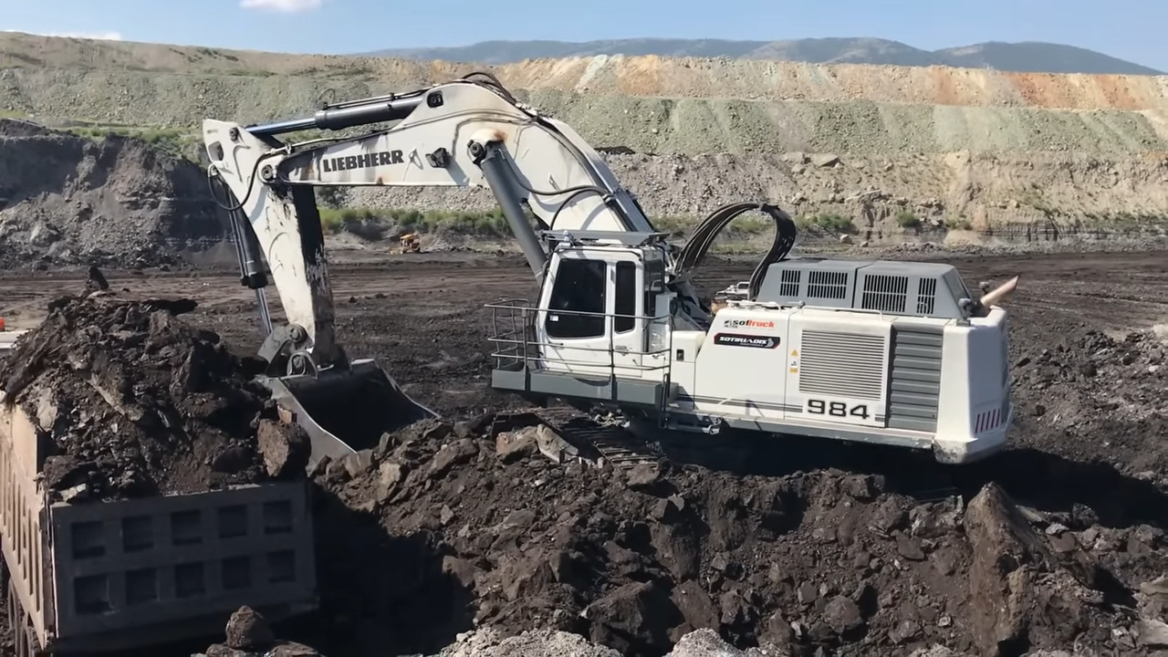 A large white excavator with the word Liebherr on its arm, scooping up a pile of dark soil in an open-pit mine with hills in the background
