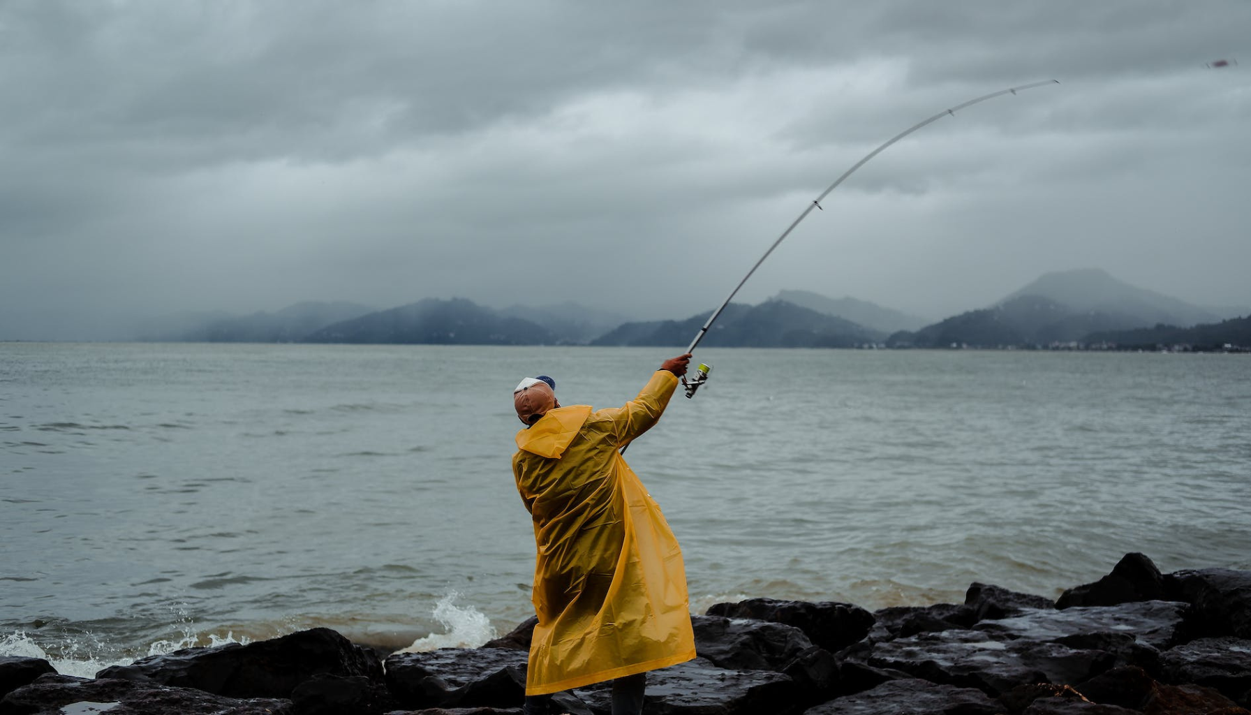 Angler in yellow raincoat casting his line from rocky terrain during gloomy weather.