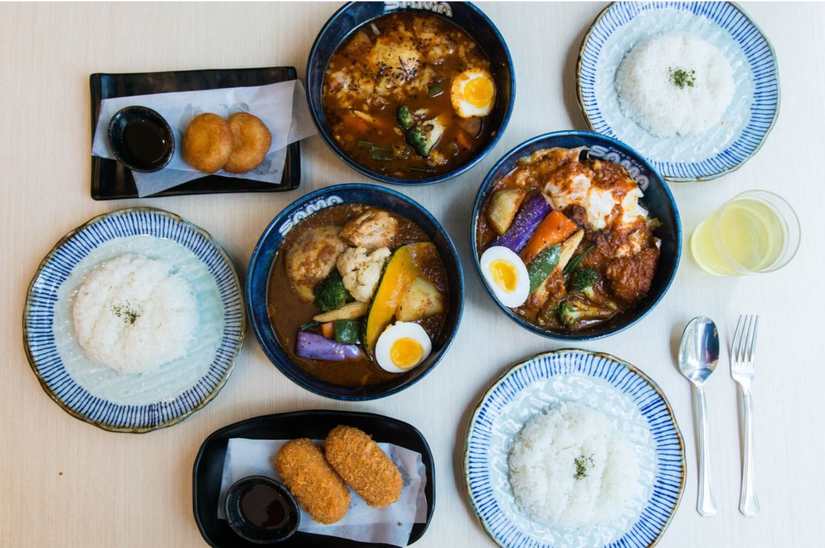 A variety of Japanese curry dishes served in bowls, accompanied by plates of white rice and garnished with vegetables and boiled eggs. To the side are fried croquettes and donuts on rectangular plates. A glass of light yellow drink and silverware are placed beside the dishes on a light wooden table, creating a vibrant, appetizing scene.