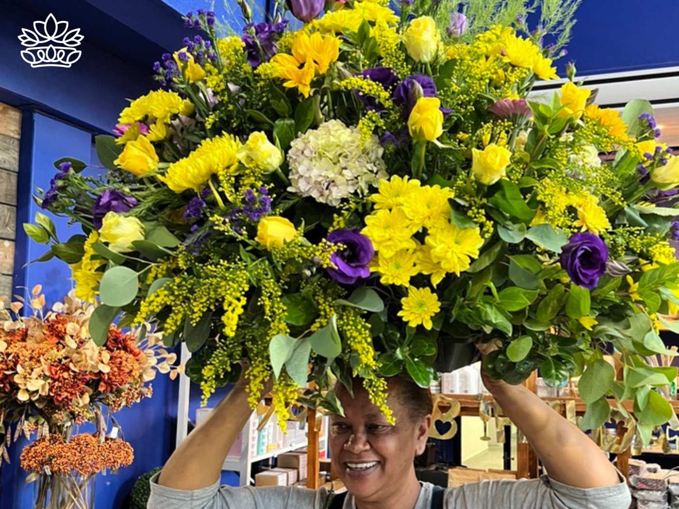 A smiling florist in a flowers shop lifting a large and vibrant flower arrangement of yellow and purple flowers overhead, showcasing an exquisite display from the Flowers By Occasion Collection at Fabulous Flowers and Gifts.