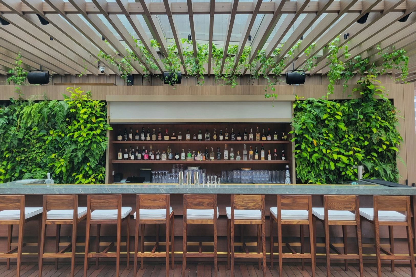Stylish outdoor bar with a modern wood pergola and lush green wall. Wooden barstools line the counter, and bottles are neatly arranged on shelves.
