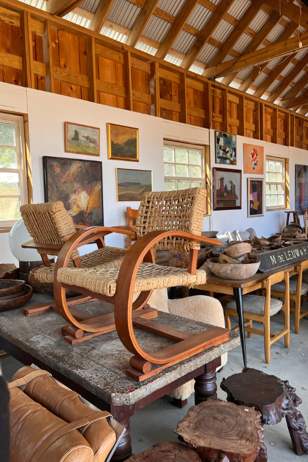 Mid-century wooden lounge chairs displayed atop a table inside a barn showroom filled with paintings, ceramics, and antique furnishings.