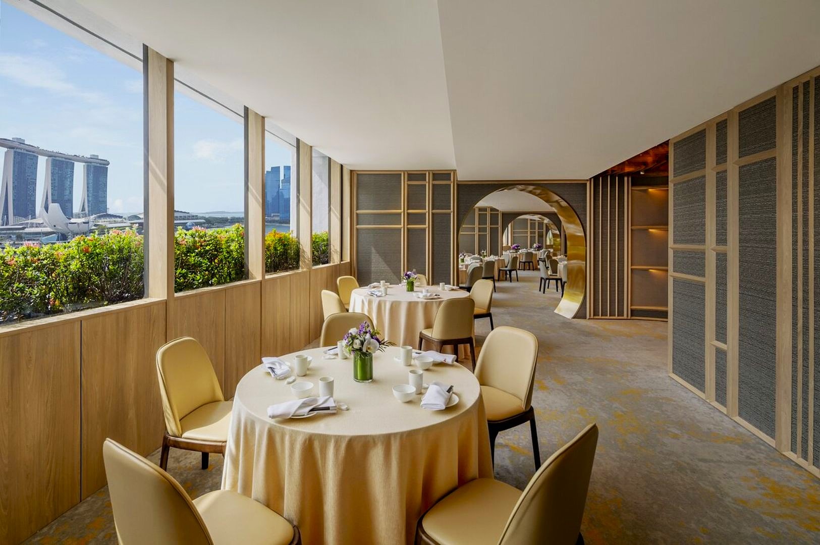 Elegant dining room with round tables set for four, white tablecloths, and floral centerpieces. Large windows reveal a cityscape. Bright and airy ambiance.