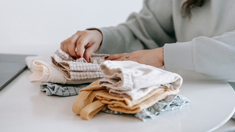 A person sitting at a table with a laptop and a pile of baby clothes