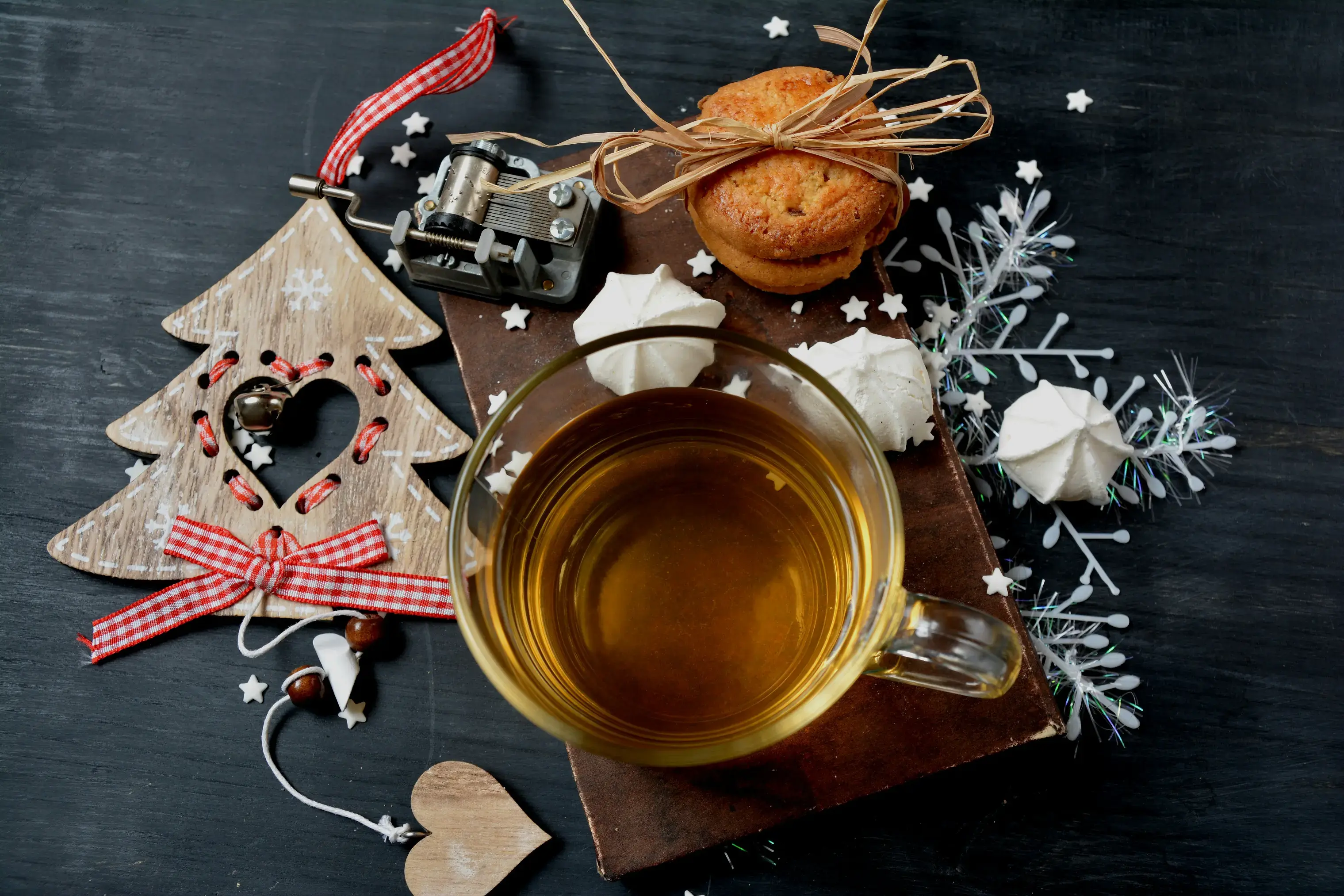 A clear glass mug filled with amber tea rests on a vintage book, surrounded by festive ornaments and sweet treats. The winter-themed flat lay is accented by a wooden Christmas tree decoration, a stack of cookies tied with twine, and delicate meringue kisses.