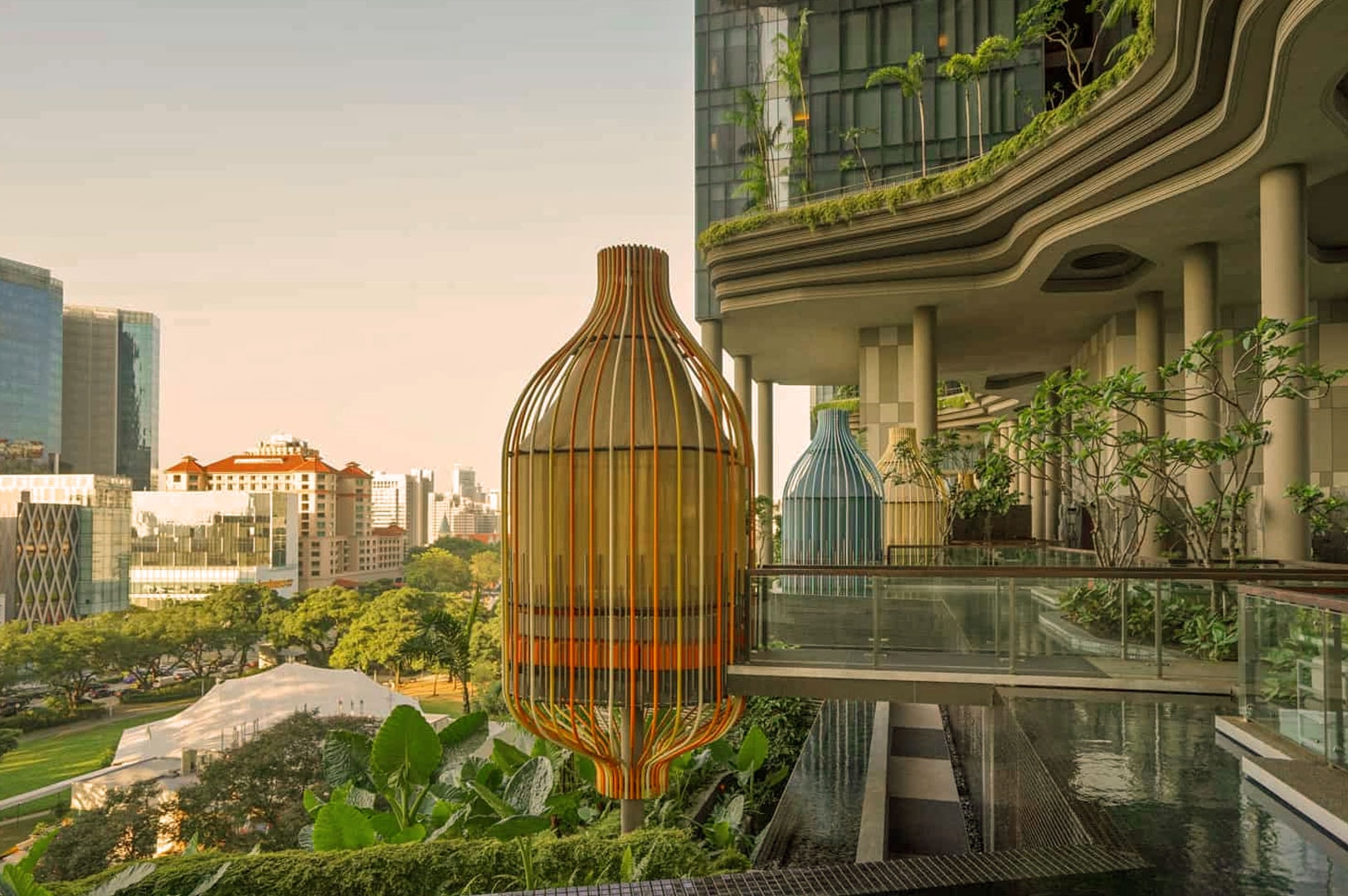 Modern urban oasis with large birdcage sculpture on a terrace. Lush greenery, reflective water features, and city skyline at sunset create a tranquil scene.