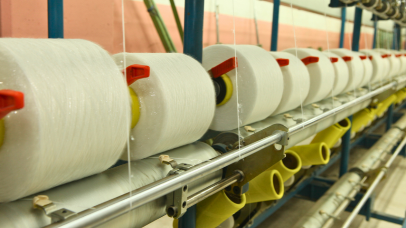 A row of large white spools of thread, contributing to the microplastic crisis, are wound onto bobbins on a machine in a factory.