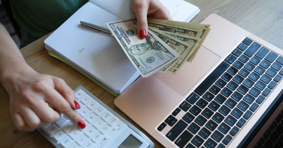 Person holding cash while calculating OnlyFans tax deductibles on a laptop, notebook, and calculator.