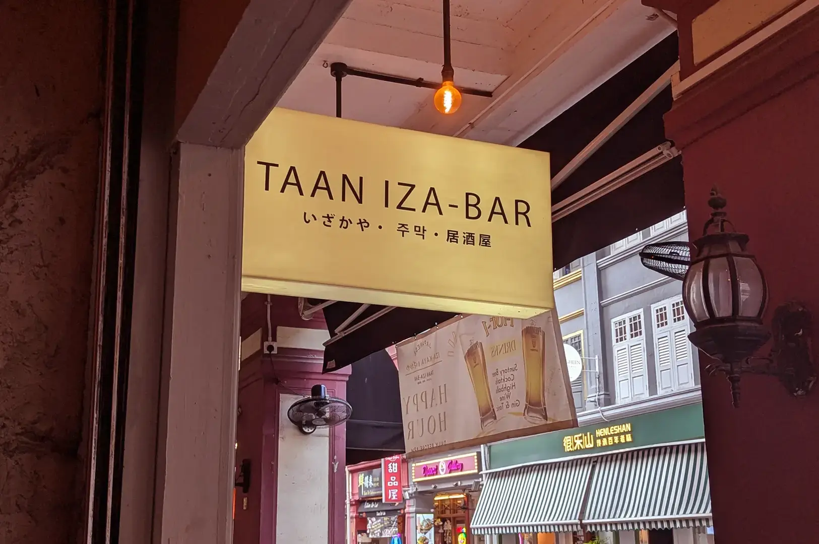 TAAN IZA-BAR entrance sign above a shophouse doorway, with Japanese text and neighboring storefronts visible across the street.
