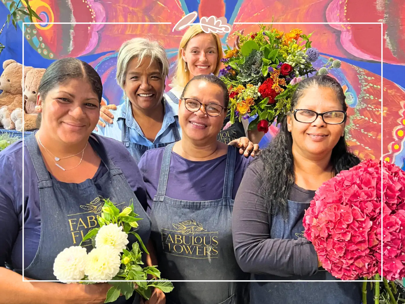 A joyful group of Fabulous Flowers staff members in aprons, holding fresh flower arrangements and smiling in front of a vibrant, colorful mural. Fabulous Flowers and Gifts.