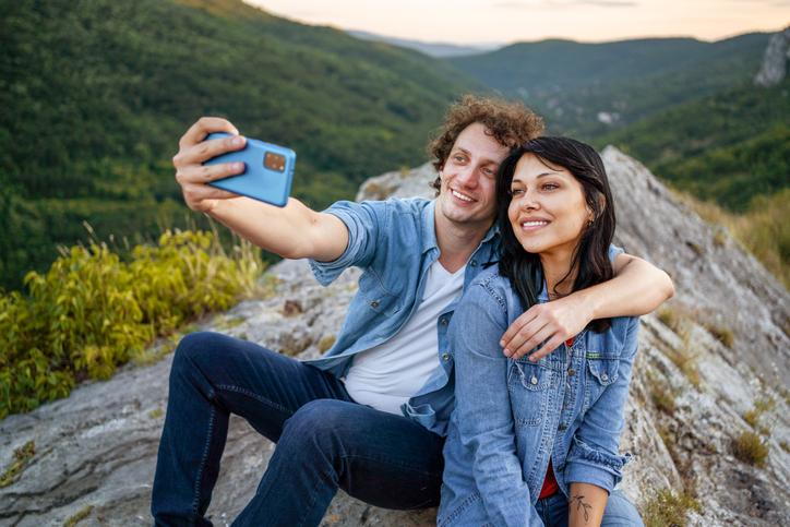 Cheerful young couple sitting on rock snapping a selfie of the mountains behind them.  