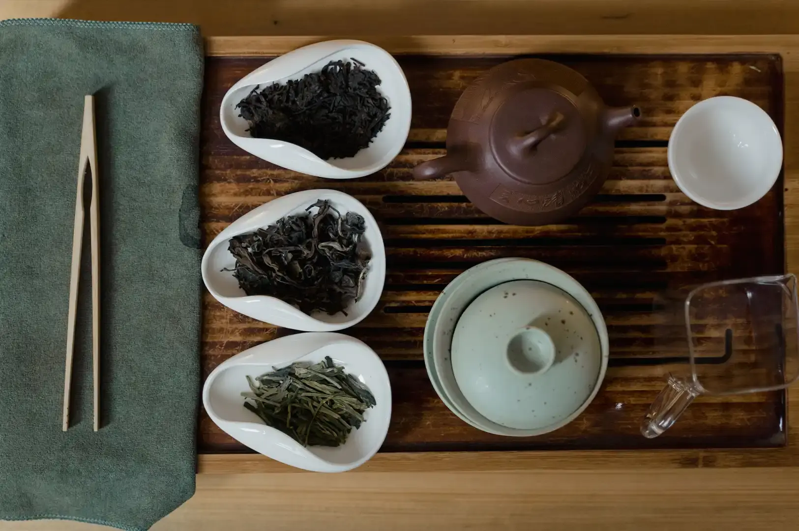 A top-down, balanced view of a traditional tea service set on a wooden tray, featuring three white, teardrop-shaped bowls filled with different loose-leaf teas, alongside a brown clay teapot, a white cup, and a pair of wooden tongs resting on a green cloth.