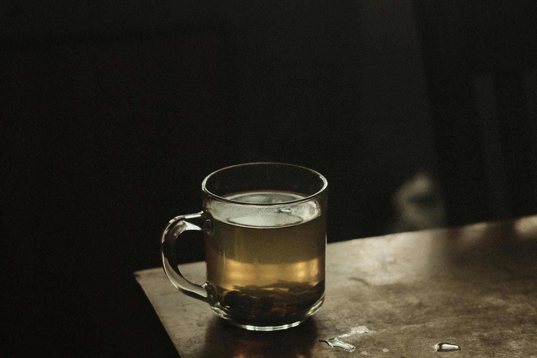 A single glass mug filled with golden tea rests on the edge of a weathered wooden table against a dark, moody background. Small droplets of water are scattered on the table's surface, catching the light alongside the tea leaves settled at the bottom of the glass.