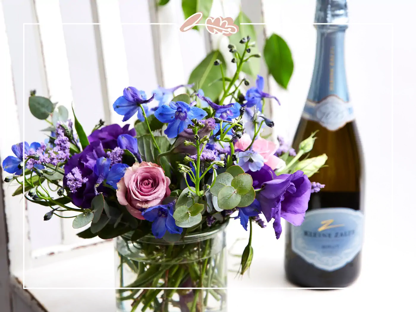 A beautiful bouquet of mixed purple and pink flowers in a glass vase, accompanied by a bottle of Kleine Zalze sparkling wine. Fabulous Flowers and Gifts.