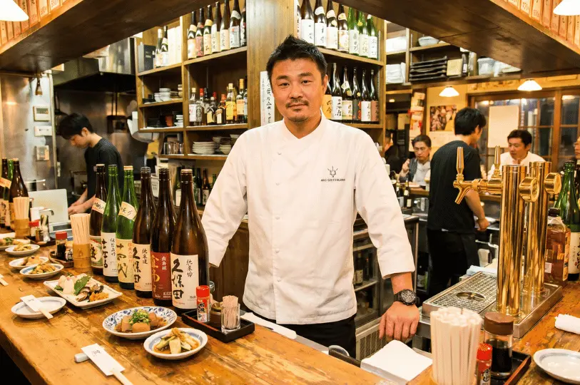 A confident male master chef in a crisp white uniform stands behind a wooden bar counter, flanked by large bottles of premium sake and small plates of appetizers.