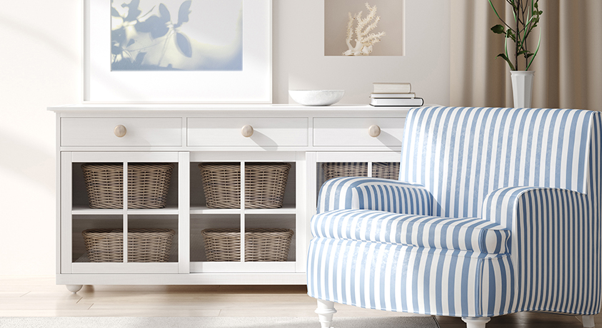 This Hamptons style room features light and bright furniture and décor, with a blue and white striped roll armchair as the main focus to contrast the off-white walls and light wooden floors. A white sideboard displays multiple wicker baskets through exposed doors. A stack of neutral books, a photo frame, an indoor plant and a piece of coral help tie the room together.