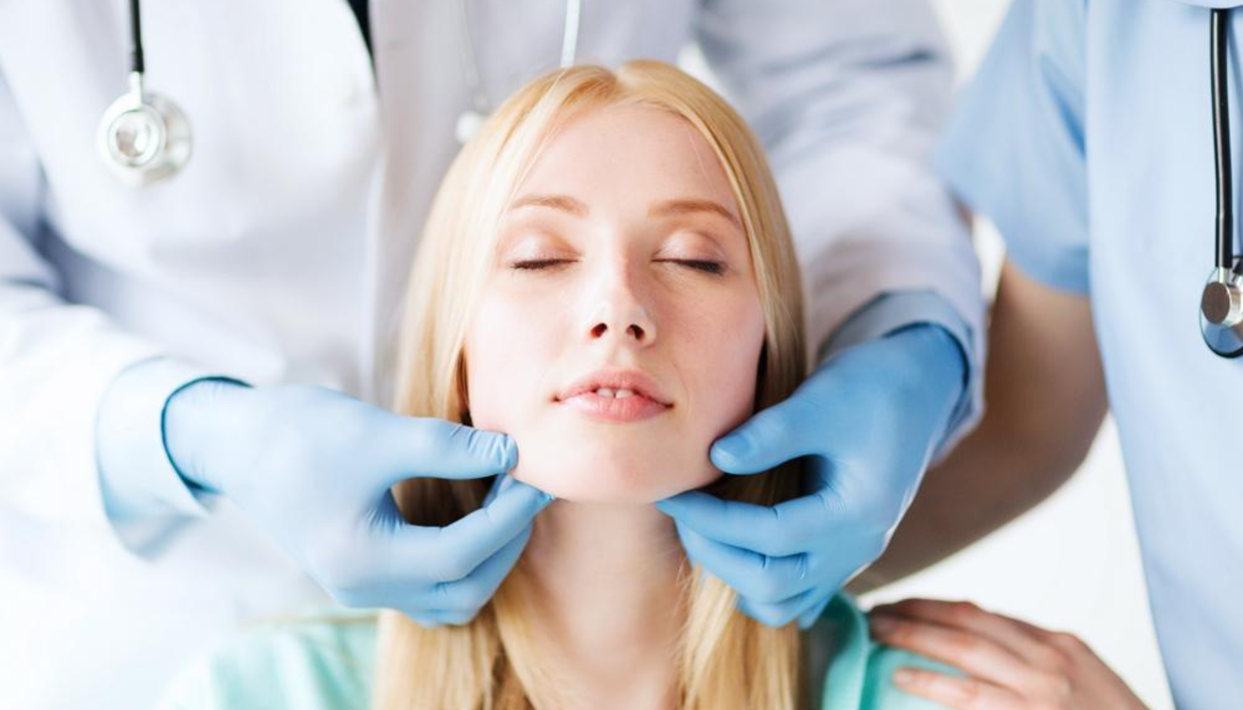 Lege undersøker kvinnens kjeve med hanskekledde hender mens pasienten lukker øynene og holder hodet stille.