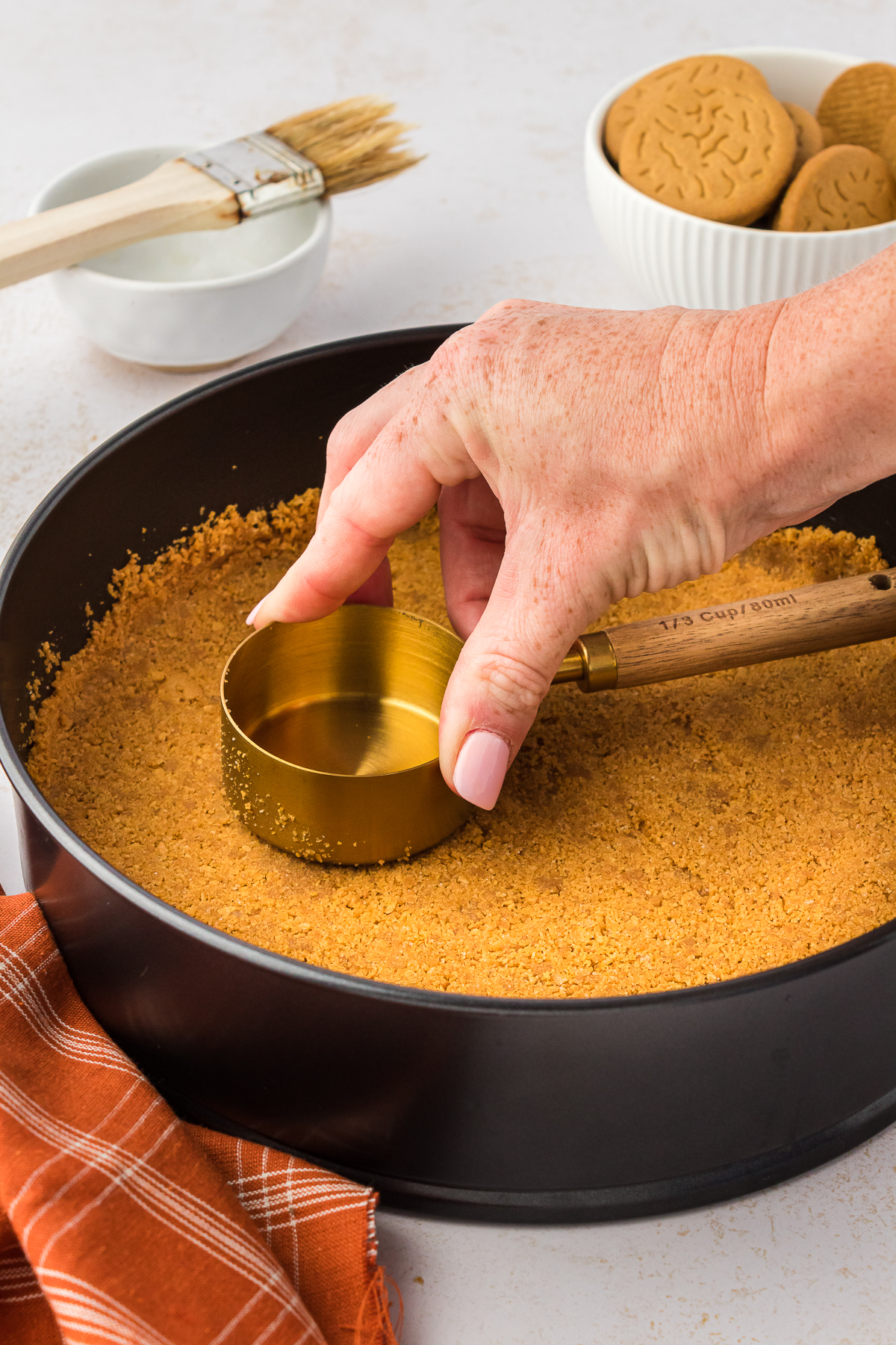 hand pressing a gingersnap crust into a 9 inch springform pan with a measuring cup