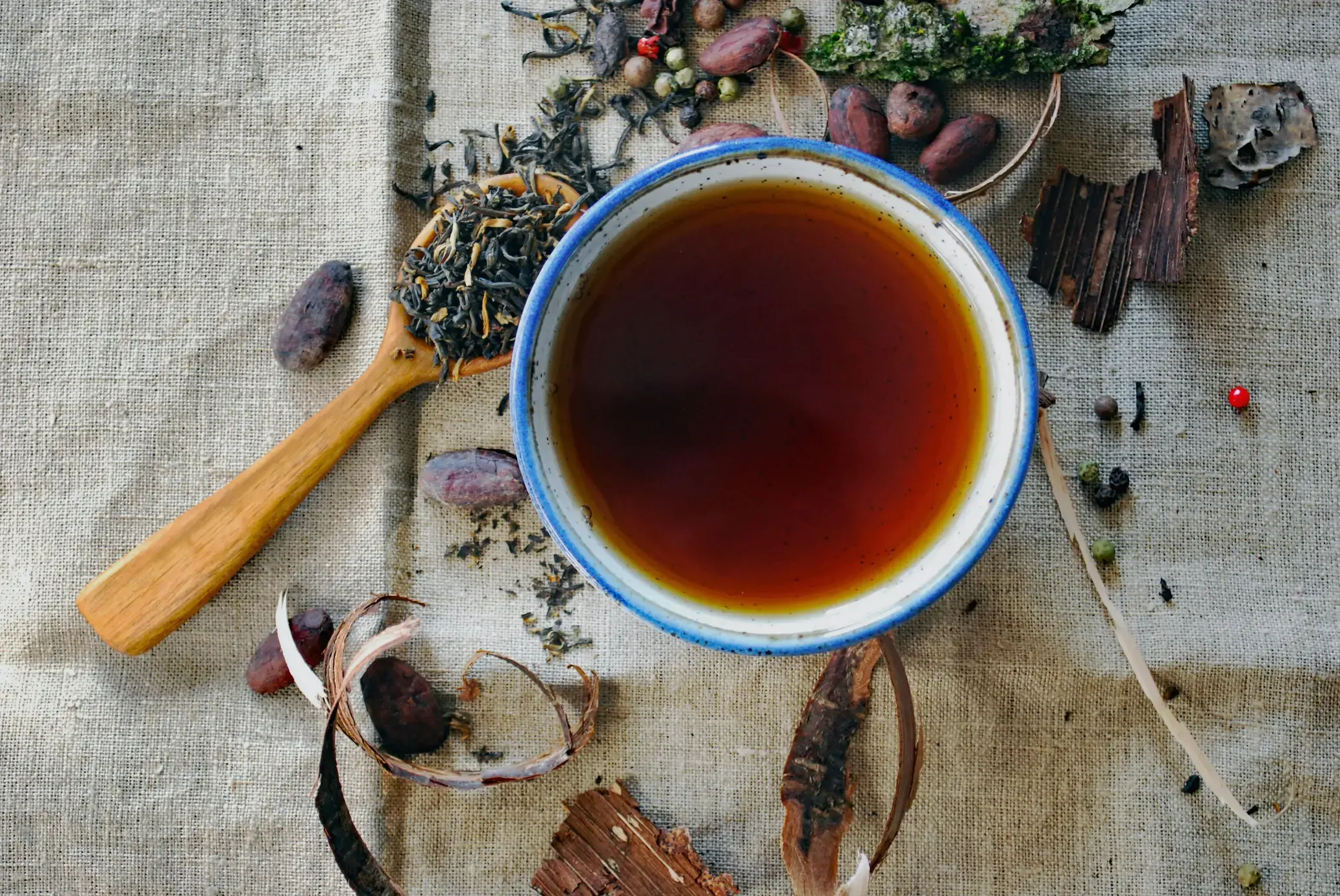 A top-down view shows a blue-rimmed ceramic cup filled with dark tea, surrounded by loose tea leaves on a wooden spoon and scattered cocoa beans. The composition is set against a textured linen backdrop featuring bits of dried bark and colorful peppercorns.