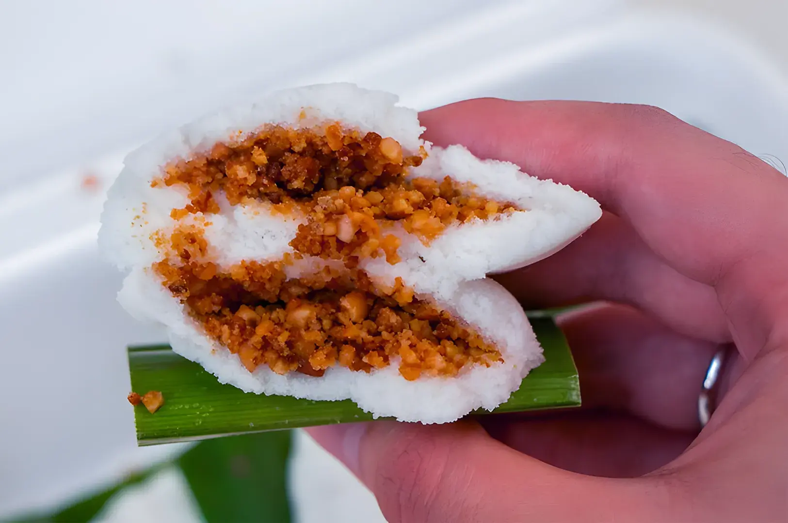 A hand holds a white, fluffy rice cake filled with a spicy, orange-colored peanut mix, resting on a small green leaf. The setting is casual and inviting.