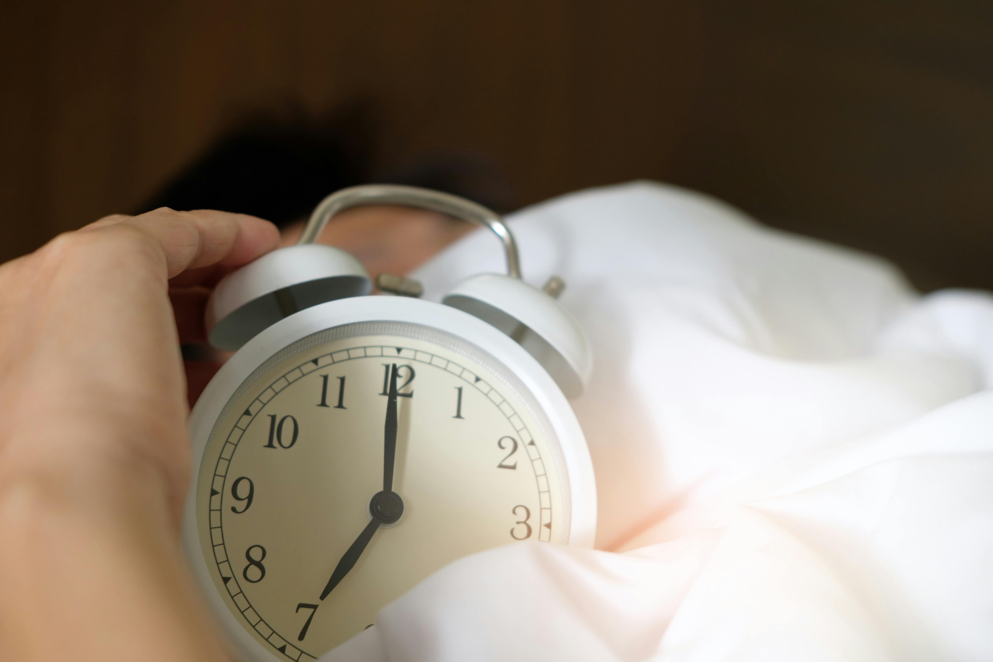 Close-up of a hand holding a white analog alarm clock showing 7:00, resting on a bed with white sheets, suggesting early morning wake-up time.