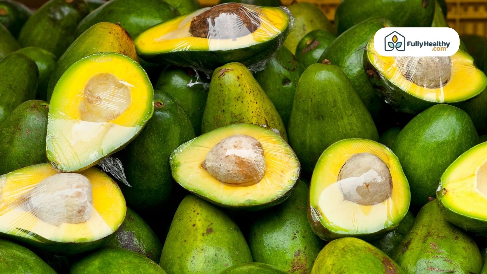 Halved avocados wrapped in plastic on top of green avocados