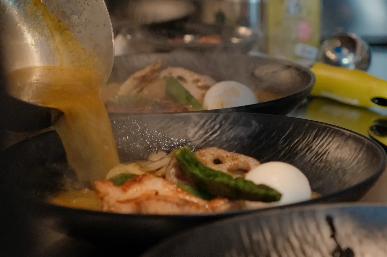 Hot broth is poured into a black bowl of noodles topped with shrimp, greens, a fish cake, and a soft-boiled egg, creating a warm, savory meal.