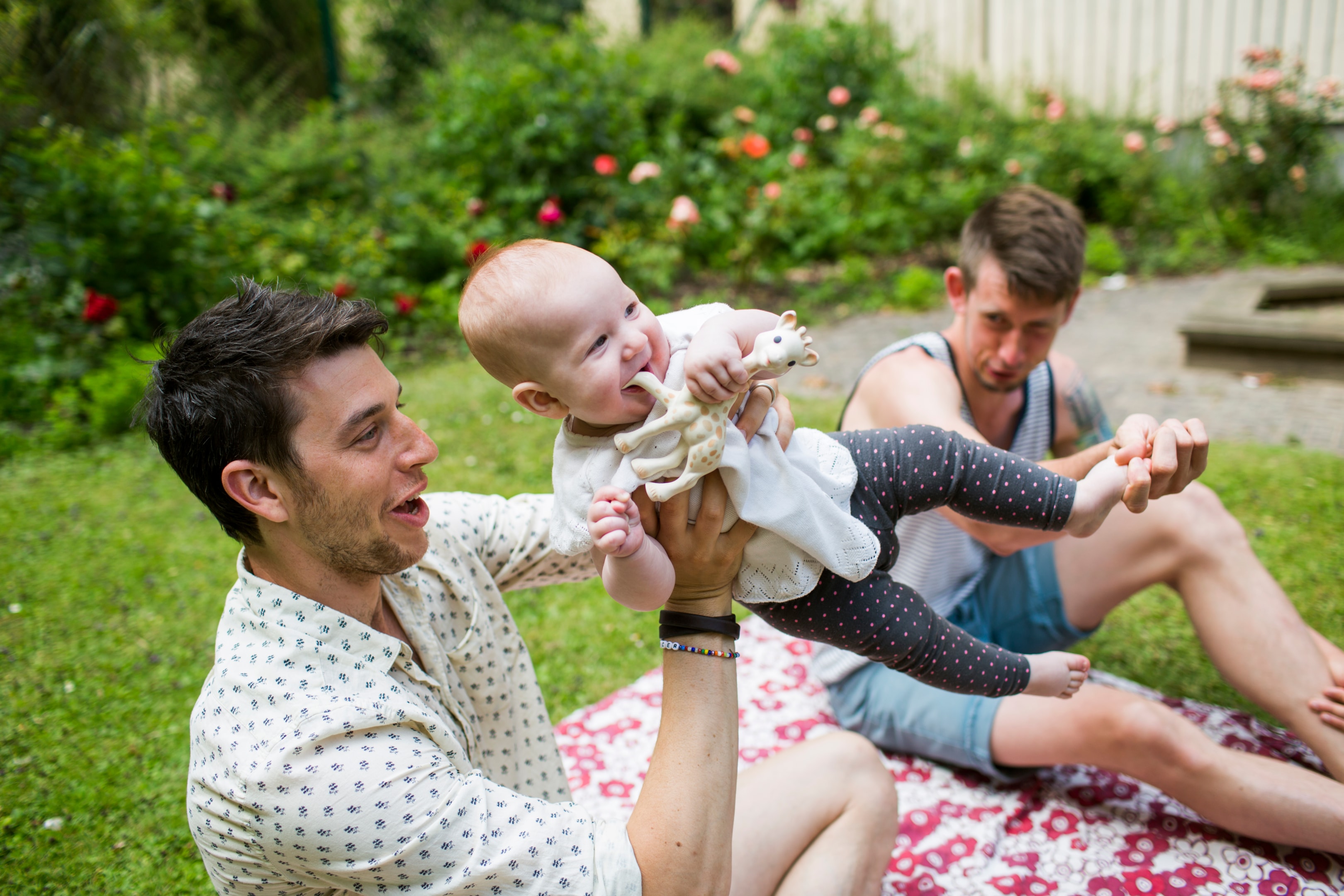 Two fathers enjoying time outdoors while holding and playing with their baby on a picnic blanket