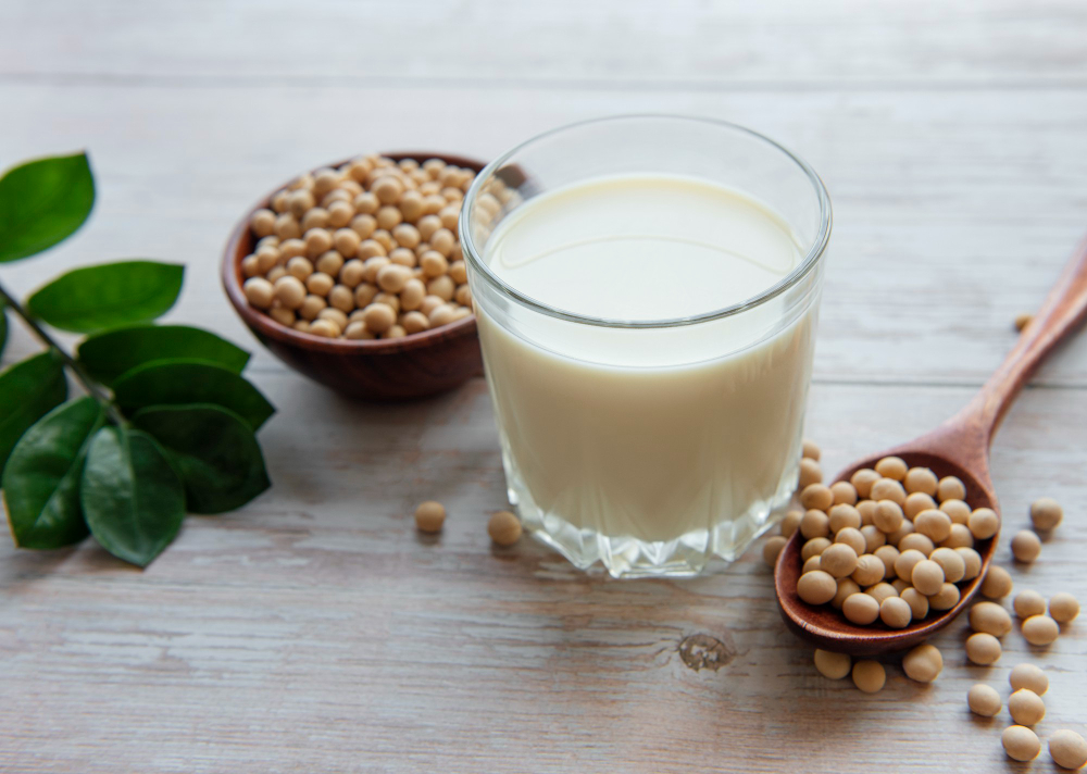 Soy milk in a glass with a bowl and wooden spoonful of soybeans around it.
