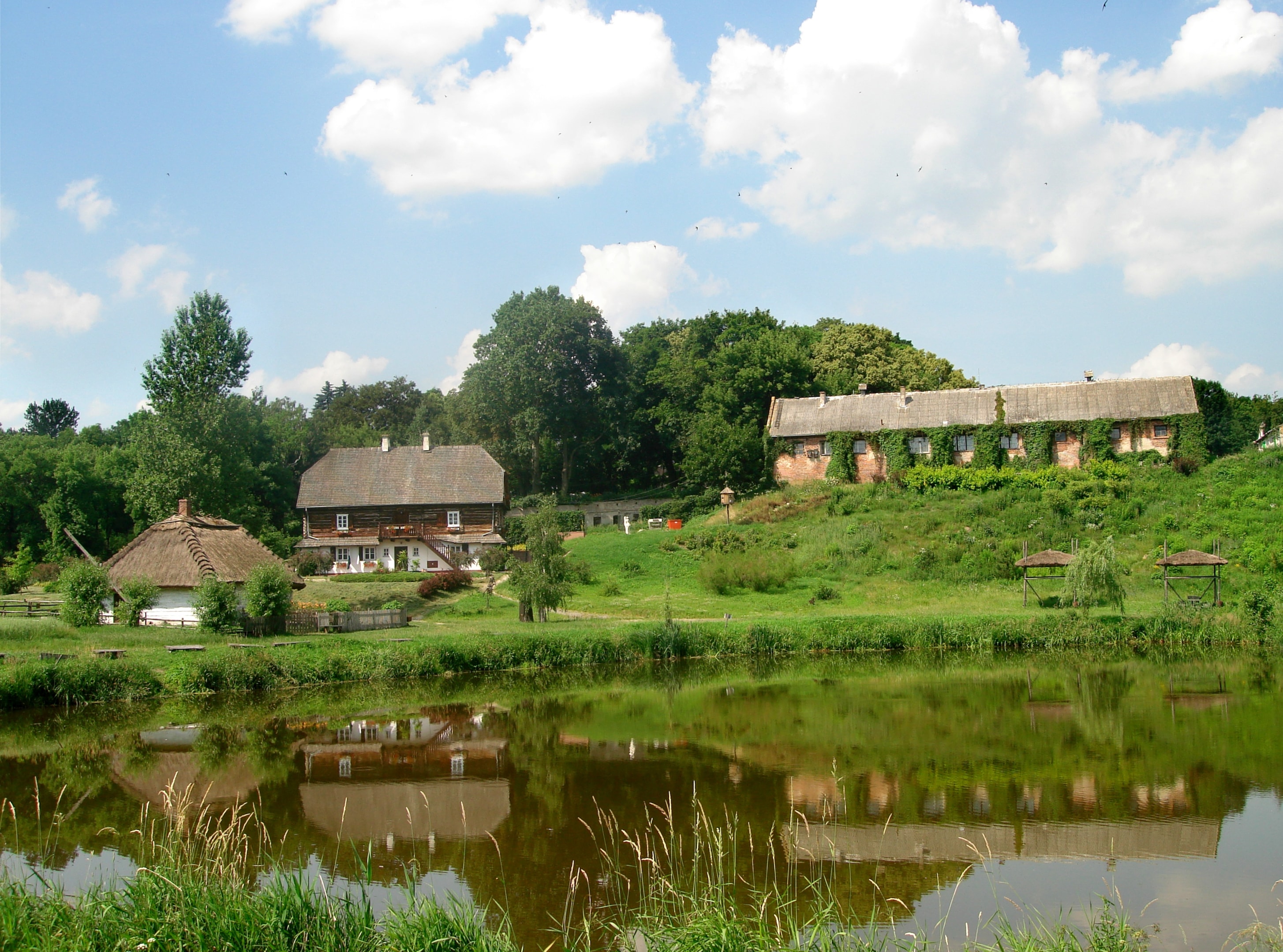 Muzeum Wsi Lubelskiej - skansen z imponującą zabudową w otoczeniu drzew i jeziora. Źródło: https://commons.wikimedia.org/wiki/File:Muzeum_Wsi_Lubelskiej_%28Skansen%29.jpg