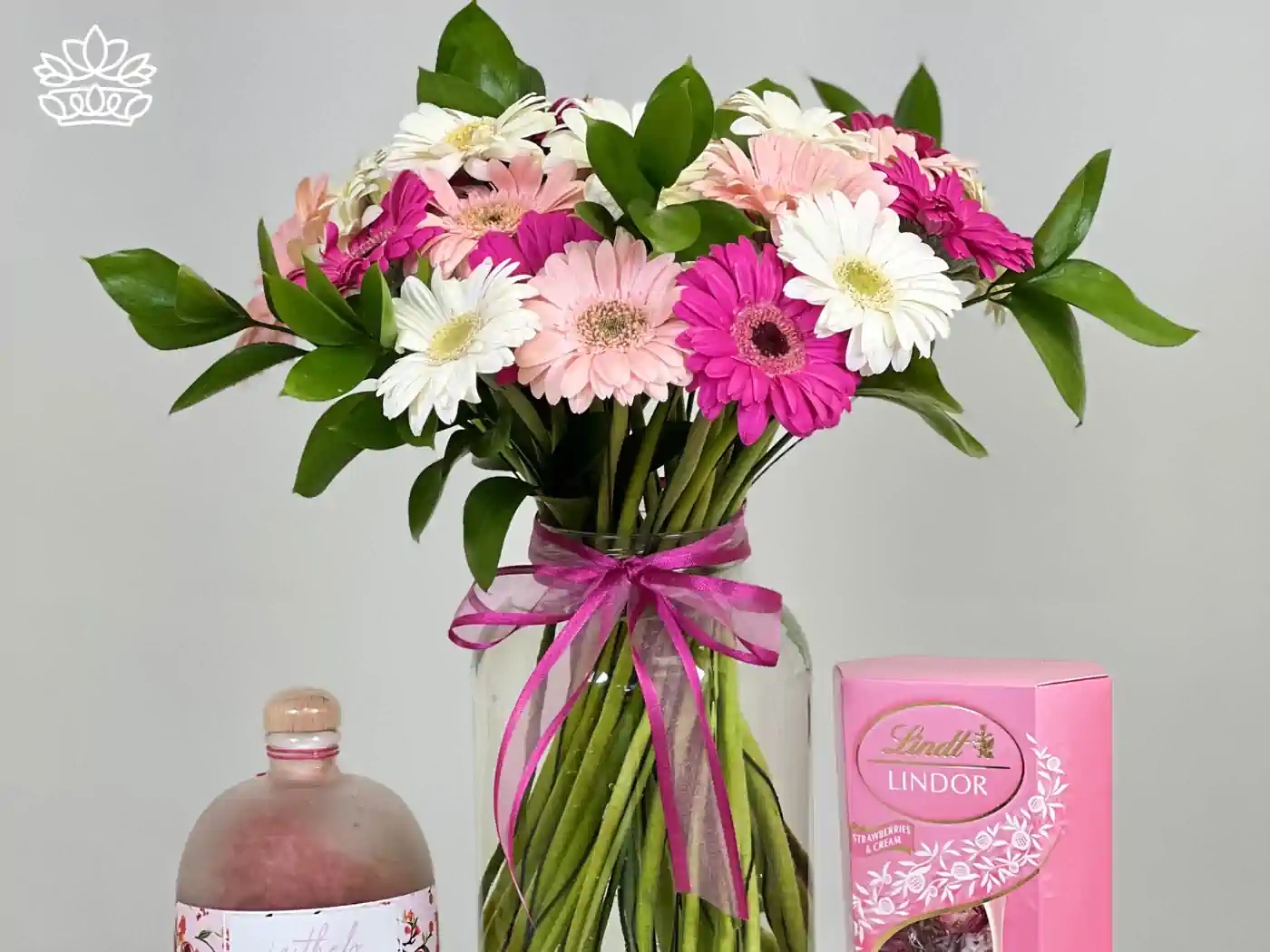 A vibrant bouquet of pink and white gerberas and daisies, elegantly tied with a pink ribbon, paired with a bottle of rose bath salts and a box of Lindt Lindor strawberries and cream chocolates. 