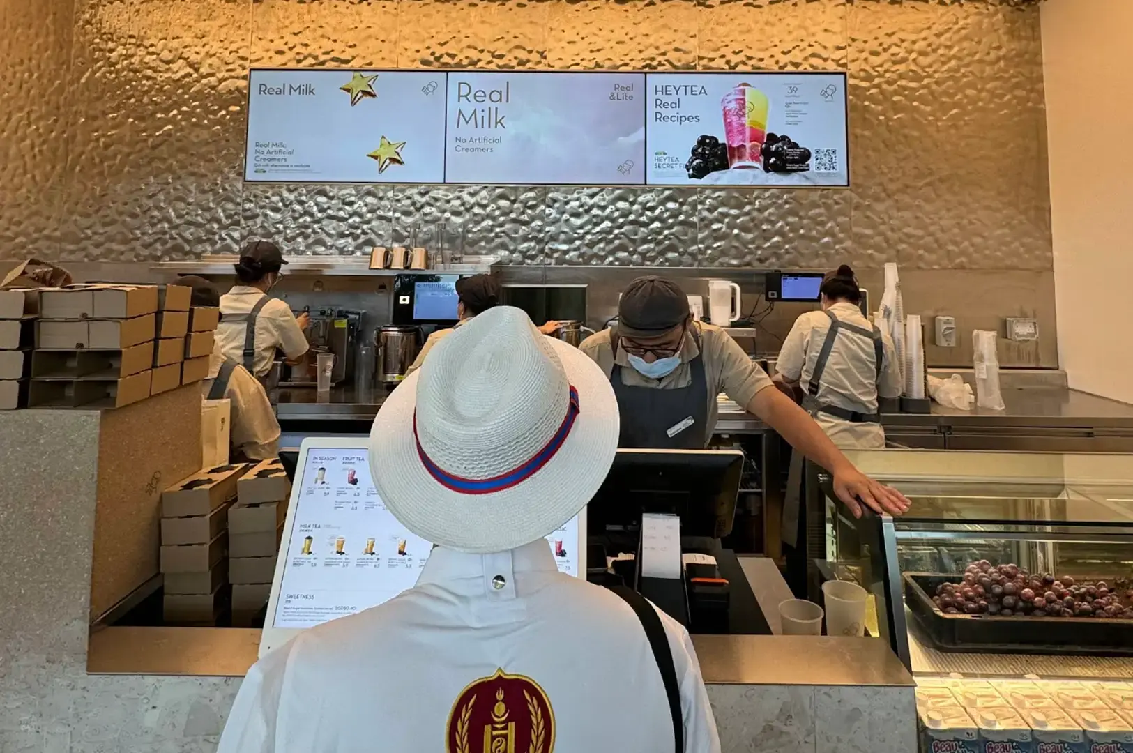 A customer wearing a white fedora hat with red and blue trim stands at the HEYTEA order counter, looking at a digital tablet menu while a staff member in a grey apron and face mask assists them beneath a hammered metallic wall and digital display screens advertising "Real Milk."