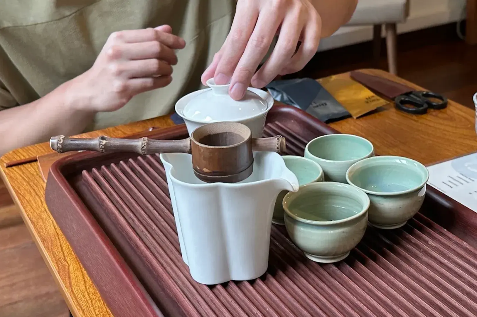 Tea service setup featuring a porcelain gaiwan, bamboo strainer, fairness cup, and celadon cups on a slatted tray.